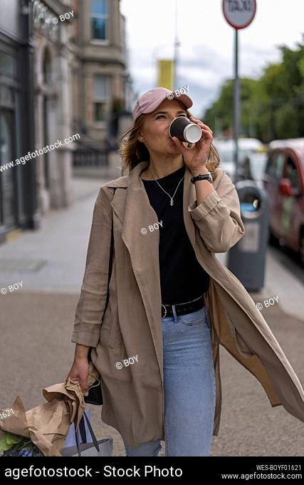 Young woman drinking coffee while carrying shopping bags walking on footpath in city