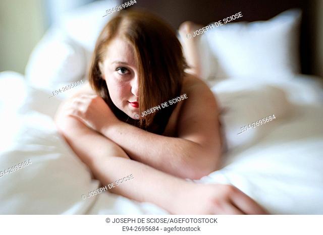 Portrait of a partially nude 24 year old woman lying on a bed and smiling at the camera