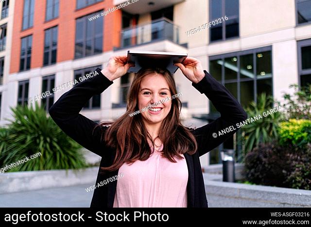 Happy businesswoman holding laptop on head in front of building