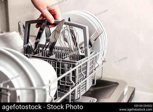 Close-up of female hand holding full cutlery basket with clean knife, fork, whisk. Loading to, empty out or unloading from open automatic dishwasher machine...