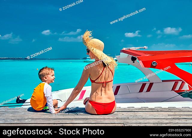Three year old toddler boy with mother sitting on wooden jetty with boat. Summer family vacation at Maldives