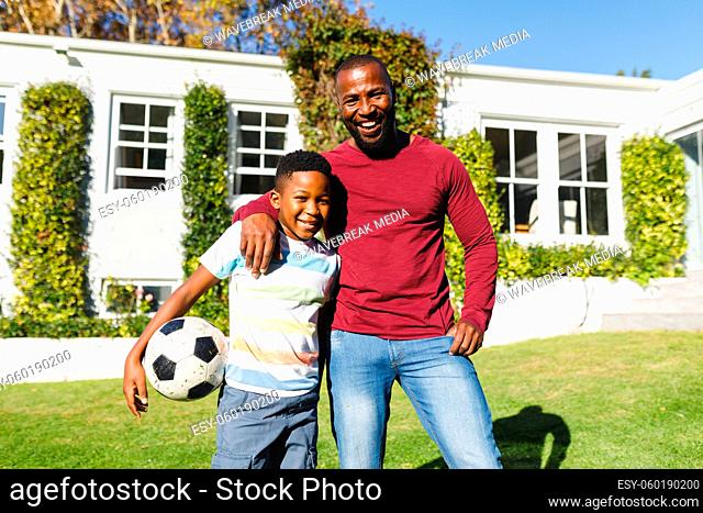 Portrait of smiling african american father with son embracing and holding football in sunny garden