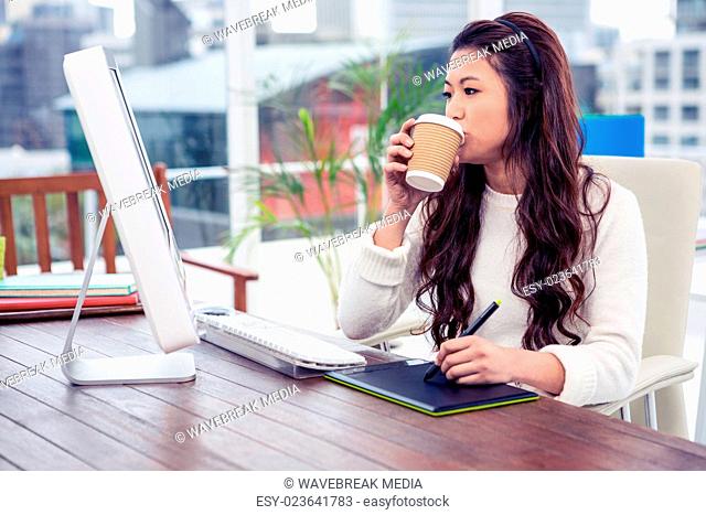 Asian woman drinking and using digital board while looking at computer monitor