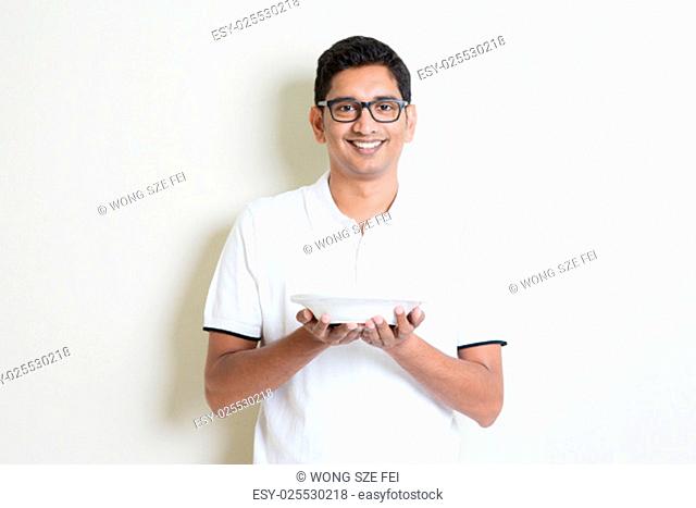 Indian guy holding a cup of coffee on hand. Asian man standing on plain background with shadow and copy space. Handsome male model