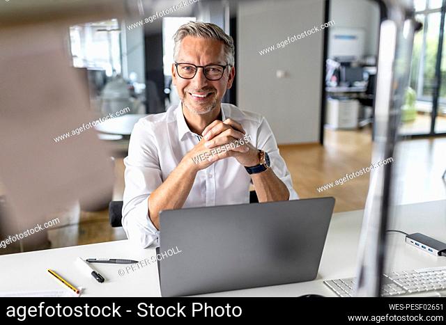 Smiling male entrepreneur with laptop sitting at desk in open plan office