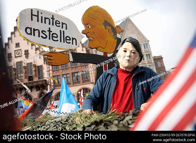 10 April 2023, Hesse, Frankfurt/Main: An activist wearing a Baerbock mask sits on a cardboard tank with a sign from Chancellor Scholz that reads ""Hinten...