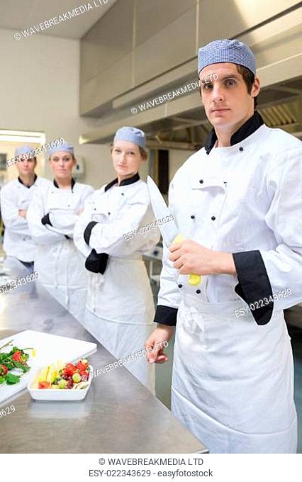 Chef looking stern holding a knife with team behind him