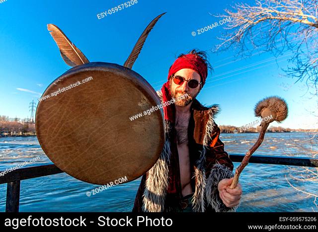 A close up view of a eagle feathers, leather drum and drumstick in the hands of a young Caucasian pagan, standing by a large river under a clear blue sky