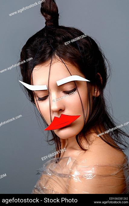 Beautiful young woman with odd fancy hairstyle and eyebrows and lips paper cutouts on face. Beauty shot on grey background. Copy space