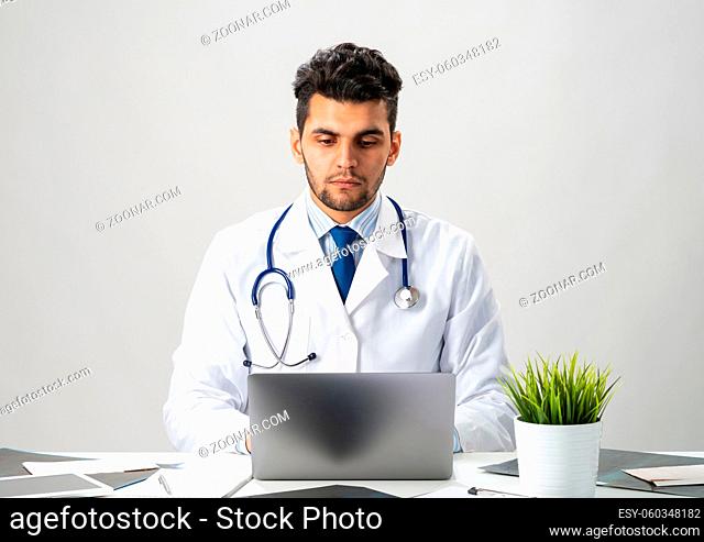 Serious arab practitioner working at computer. Handsome physician in white medical gown with stethoscope sitting at desk with laptop