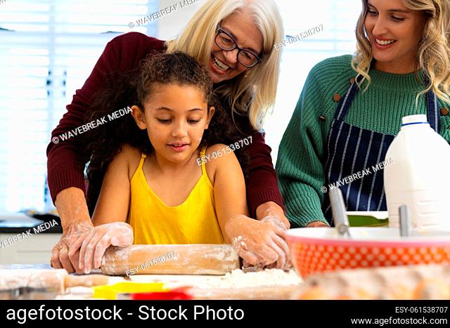 Multiracial multigeneration female family kneading dough with rolling pin on table in kitchen