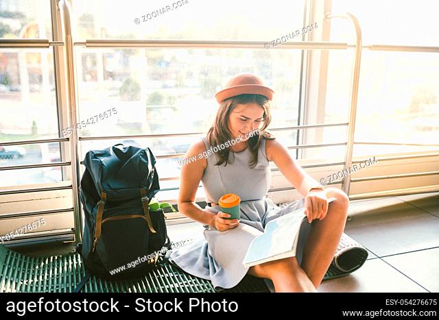 Waiting, delayed transport in the terminal of the airport or train station. Young caucasian woman in dress and hat sits on tourist rug with backpack near window...