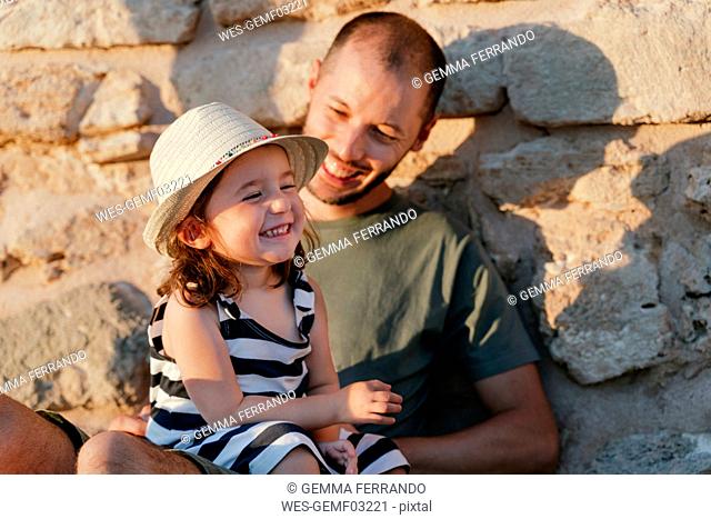 Portrait of happy little girl sitting on her father's lap at sunlight
