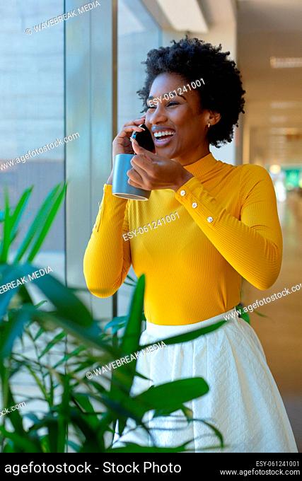 Happy african american businesswoman drinking coffee while talking on smartphone at workplace