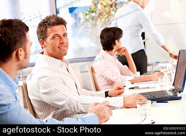 Mid-adult businessman sitting on business meeting with colleagues at office, looking at camera, smiling