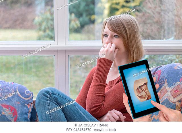 Sad young woman is sitting at home and another female hands are showing space shot of the unborn child in the tablet screen