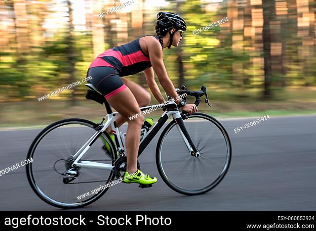 Cute sportive girl rides a bike on the road on the nature background. She wears black-pink sportswear, a stopwatch, a black helmet