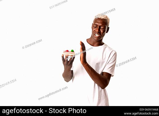 Portrait of disgusted handsome african american young blond man, unwilling eat dessert, cringe from disgusting cake, standing white background