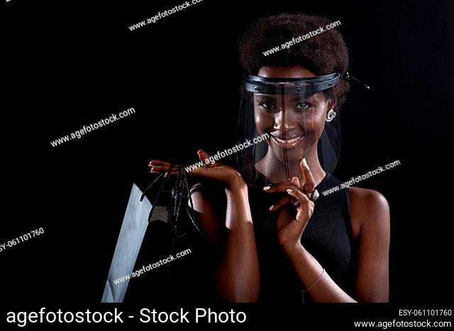 African american woman in black medical face mask and transparent protective face shield holds black shopping bags. Safety on Black Friday concept