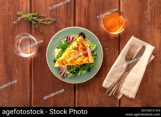 A slice of a French quiche with salmon, with green salad leaves, thyme and white wine, shot from above on a dark rustic wooden background
