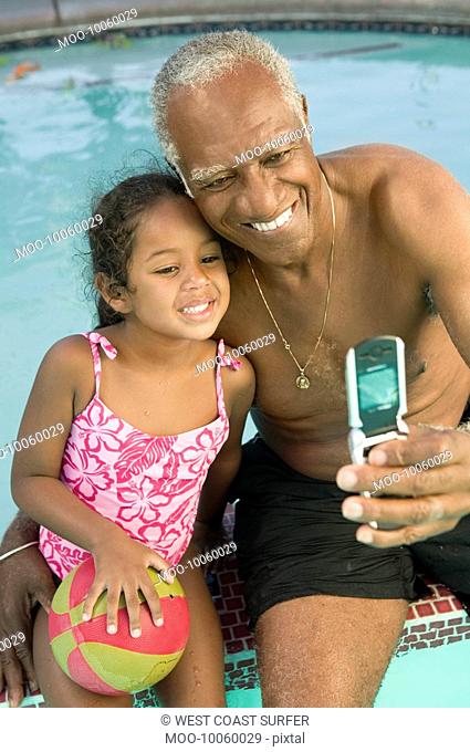 Senior man using mobile phone photographing self with granddaughter 5-6 at swimming pool