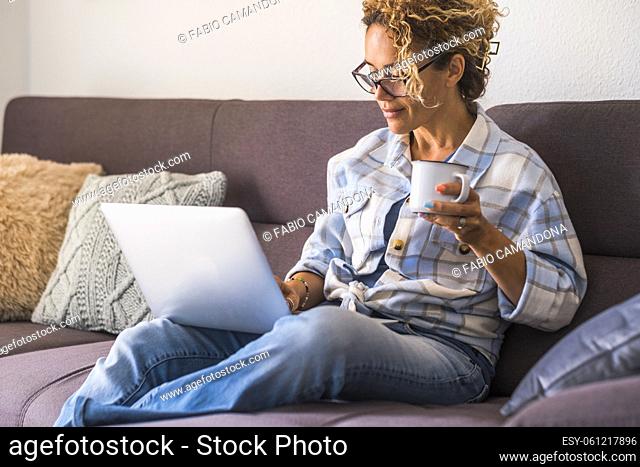 Confident happy adult young woman use laptop computer sitting on the sofa at home in relax leisure activity drinking coffee or tea and smiling - modern...