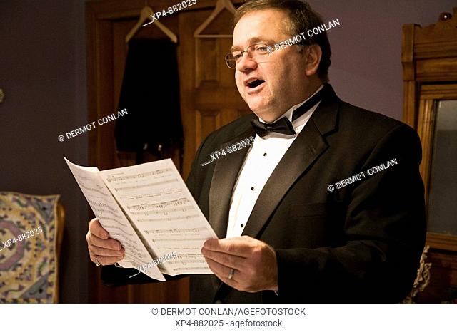 A man wearing a black tuxedo and bow tie singing from a book of sheet music in his dressing room