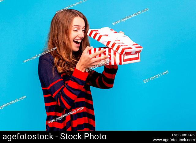 Side view portrait of woman wearing striped casual style sweater, opens gift box, looking inside with happy expression, being glad to get such present