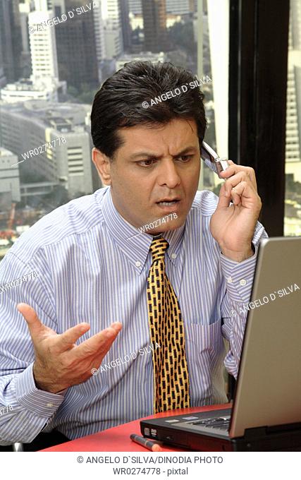 Executive looking at screen of laptop with talking on mobile showing worried expression in office at top floors of skyscraper in modern city MR687U