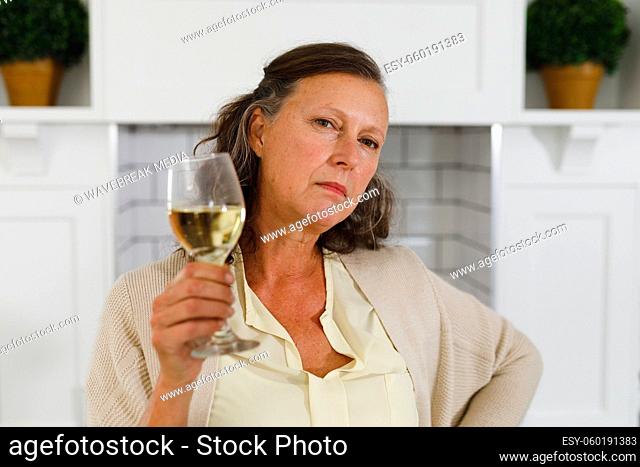 Portrait of senior caucasian woman in modern kitchen, holding glass of wine, looking to camera