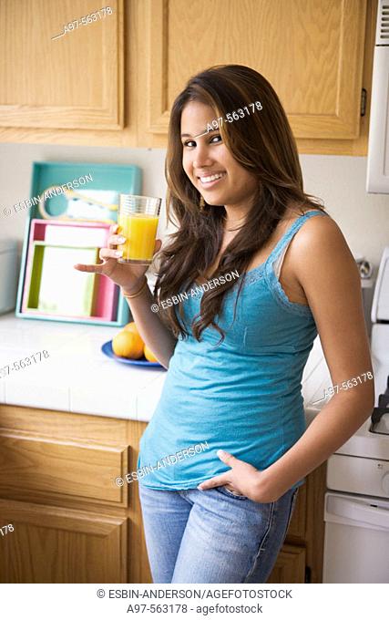 Smiling latina teen girl in blue tank top holding a glass of orange juice while standing in kitchen