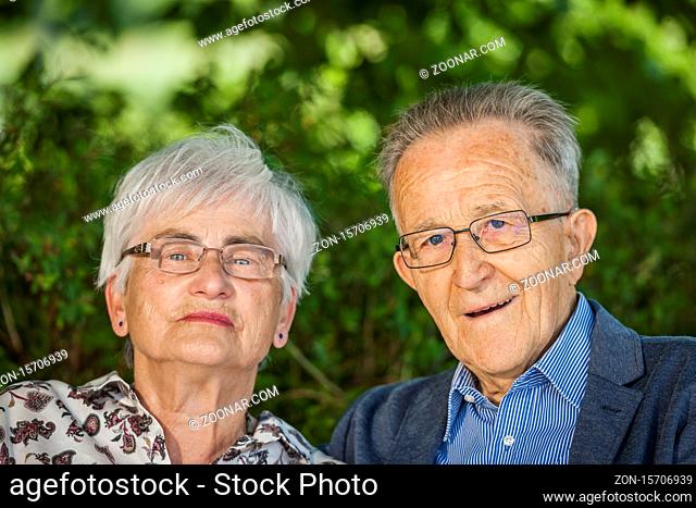 Kopf-und-Schulter-Porträt eines älteren Rentner Paares mit Brillen vor grünen Laubblättern im Sommer freudig in die Kamera blickend