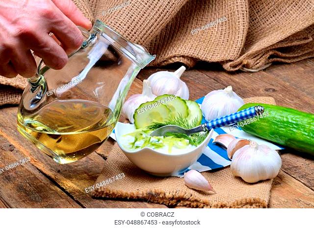 Man pours oil into Tzatziki - traditional Greek dressing or dip sauce, garlic, lemon, dill, cucumber, jug with oil, blue spoon and decoration in background