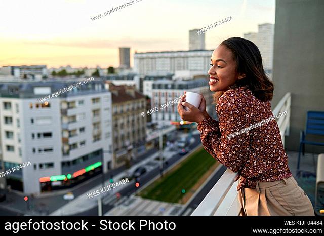 Smiling young woman holding coffee cup in balcony at sunset