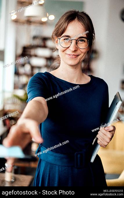Smiling young female freelancer holding laptop giving card in cafe