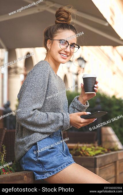 Young smiling woman in glasses with mobile phone and takeaway coffee cup standing at outdoors cafe