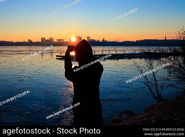 dpatop - 14 December 2022, Hamburg: A jogger takes a photo of the sunrise over the Alster at the Rabenstraße jetty with her smartphone