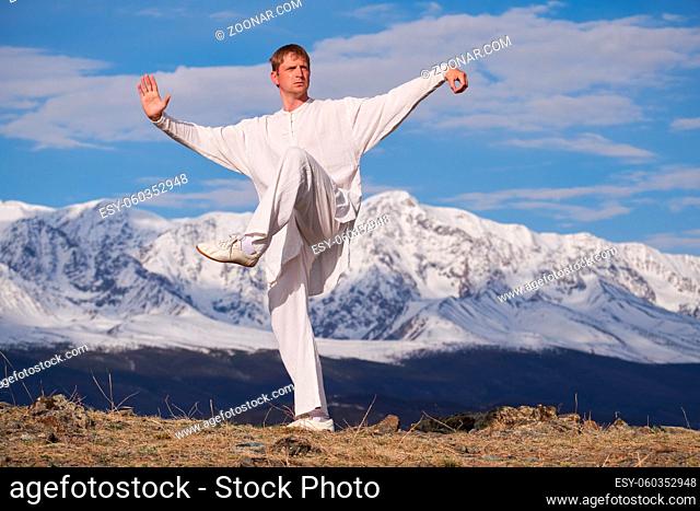 Wushu master in a white sports uniform training on the hill. Kungfu champion trains maritial arts in nature on background of snowy mountains