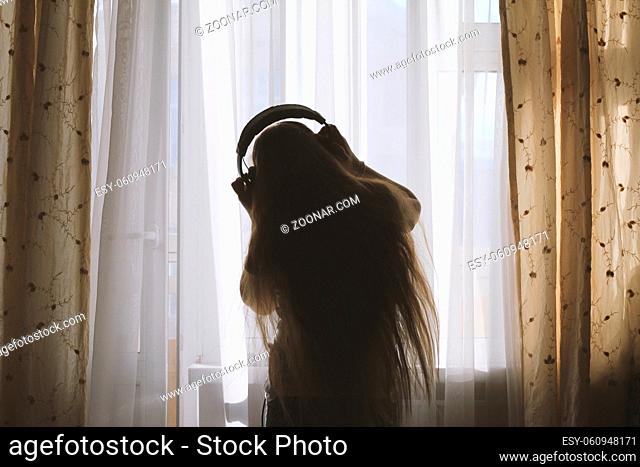 Silhouette of teenager girl in headphones listening to music and dancing in front of the window, concept of youth leisure