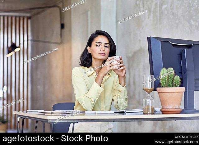 Thoughtful businesswoman holding coffee mug while sitting at desk in office