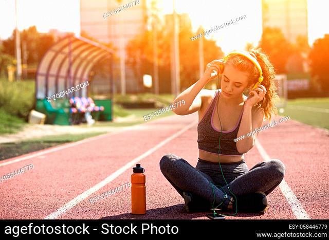 Girl is Having Rest After Training. Beautiful Skinny Blond in Sports Top is Listening to Music with Eyes Closed. Woman is Sitting with Crossed Legs on Running...
