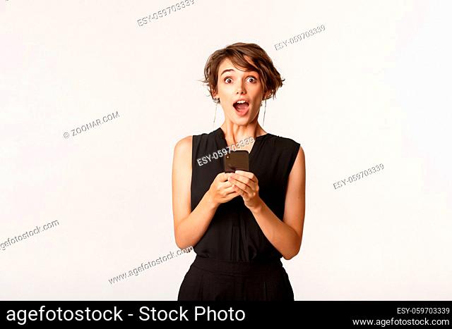 Amazed beautiful female in elegant clothes holding smartphone, open mouth surprised after reading message, standing white background