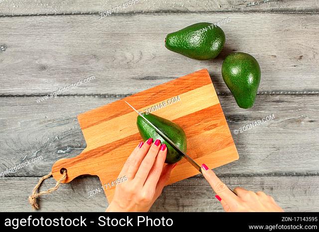 Tabletop view, young woman cutting whole avocado on chopping board