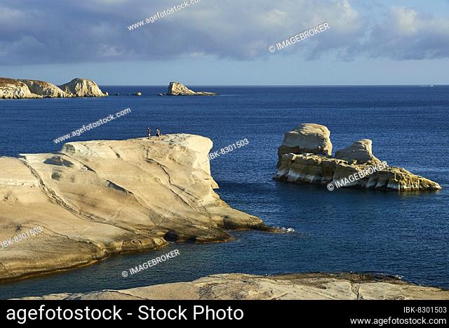 Morning light, tuff rock, tuff rocks, offshore islets, 2 people on rocks, calm dark blue sea, light blue sky, white clouds, Sarakiniko Beach, Milos Island