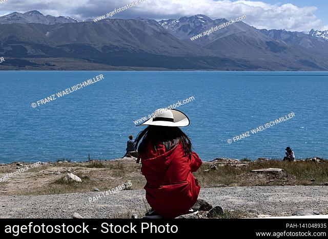 Lake Pukaki; a woman in a sun hat, photographed from behind, is sitting on the lake shore; on January 5th, 2019 near Twizel in the Canterbury region on the...