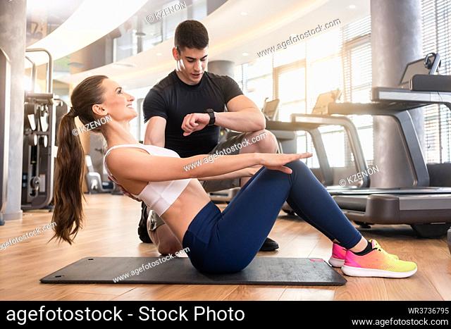 Experienced personal trainer timing and guiding young fit woman during isometric exercise for abdominal muscles in a modern fitness center