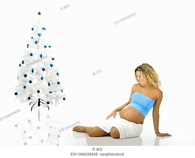 Beautiful pregnant woman seated on the floor near a white christmas tree