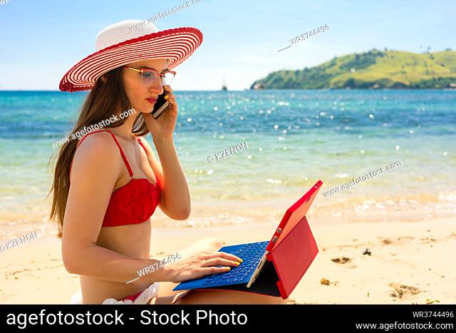 Fashionable young woman talking on mobile phone while using a tablet on the beach in a sunny day of summer in Flores Island, Indonesia