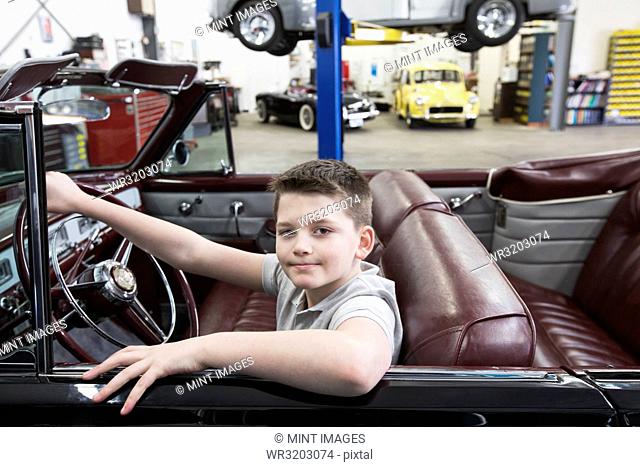 A young Caucasian boy at the wheel of an old convertible in his dad's classic car repair shop