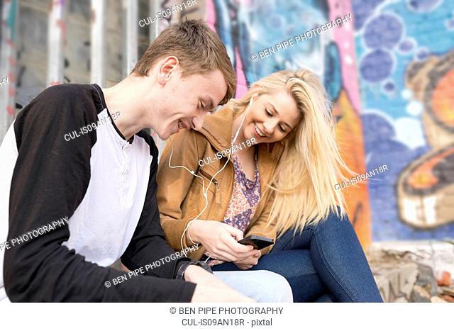 Teenage couple listening to mp3 player against wall with graffiti